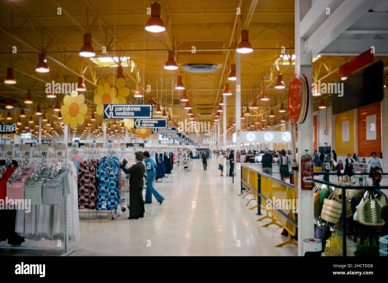 The interior of large Spanish supermarket in 1977. The vast warehouse area includes clothing, seen here left. Giant cut-out yellow flowers hang from the ceiling and the store’s colour palette emphasises bright yellow, red and orange – primary colours that were popular at that time. This image is from an amateur 35mm colour transparency in poor lighting conditions – a vintage 1970s photograph. Stock Photo