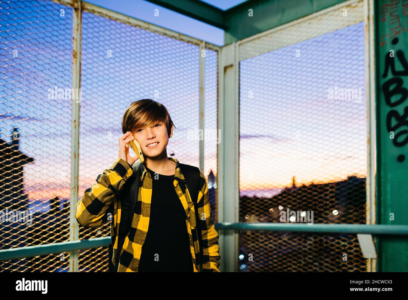 Teenager boy talking on a yellow smartphone while crossing an old metal bridge, on his way to school Stock Photo