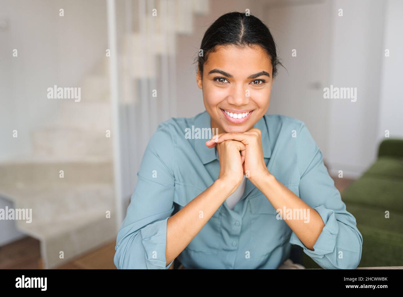Portrait of charming elegant woman with hands under chin smiling at the camera, sitting in the modern house, wearing jeans shirt Stock Photo