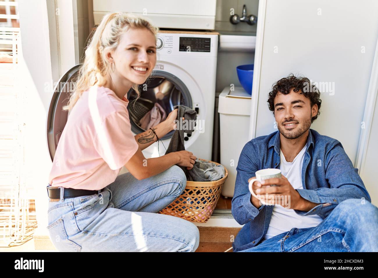 Young couple smiling happy drinking coffee while doing laundry at home ...