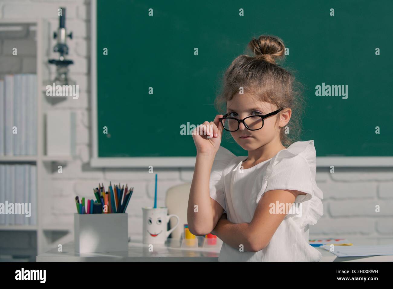 Close-up portrait of attractive small little cheerful girl sitting on table  desktop in class room indoors. Little funny school girl face. Nerd school  Stock Photo - Alamy