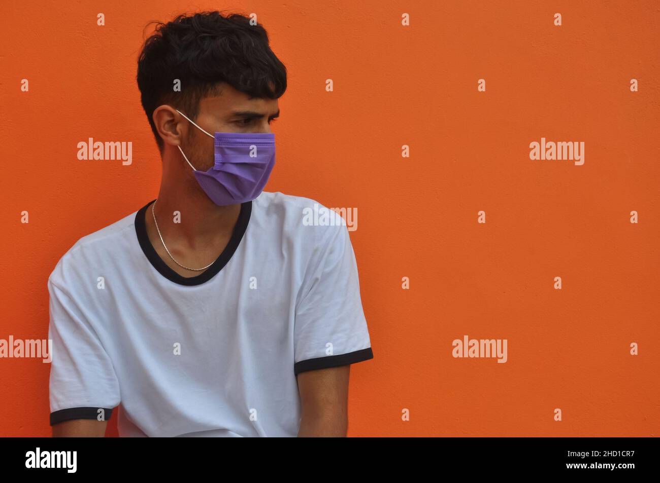 A good looking Indian young guy wearing face mask with looking sideways sitting against orange wall background with copy space Stock Photo