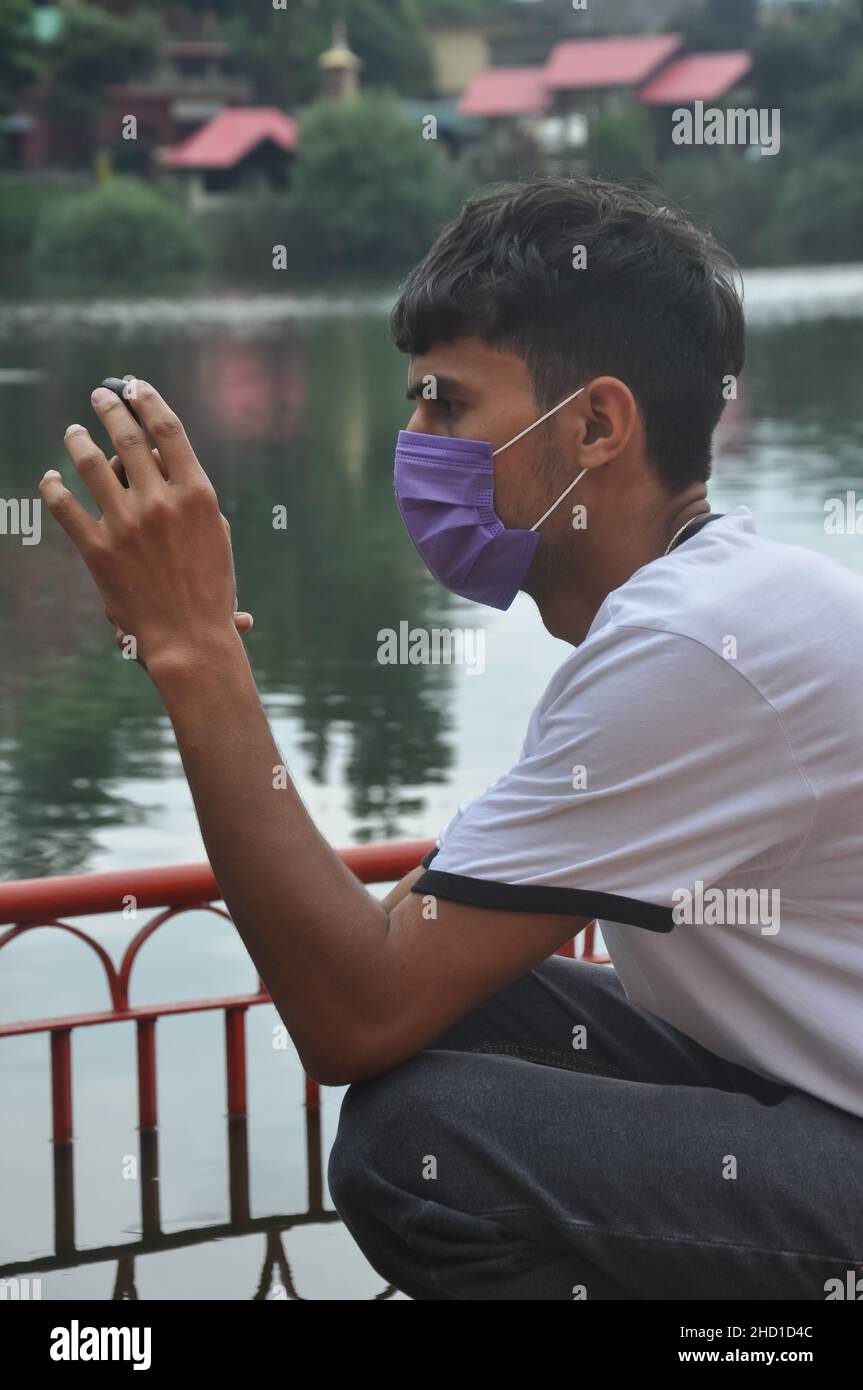 Side view of a guy using mobile phone, wearing face mask sitting beside the lake Stock Photo