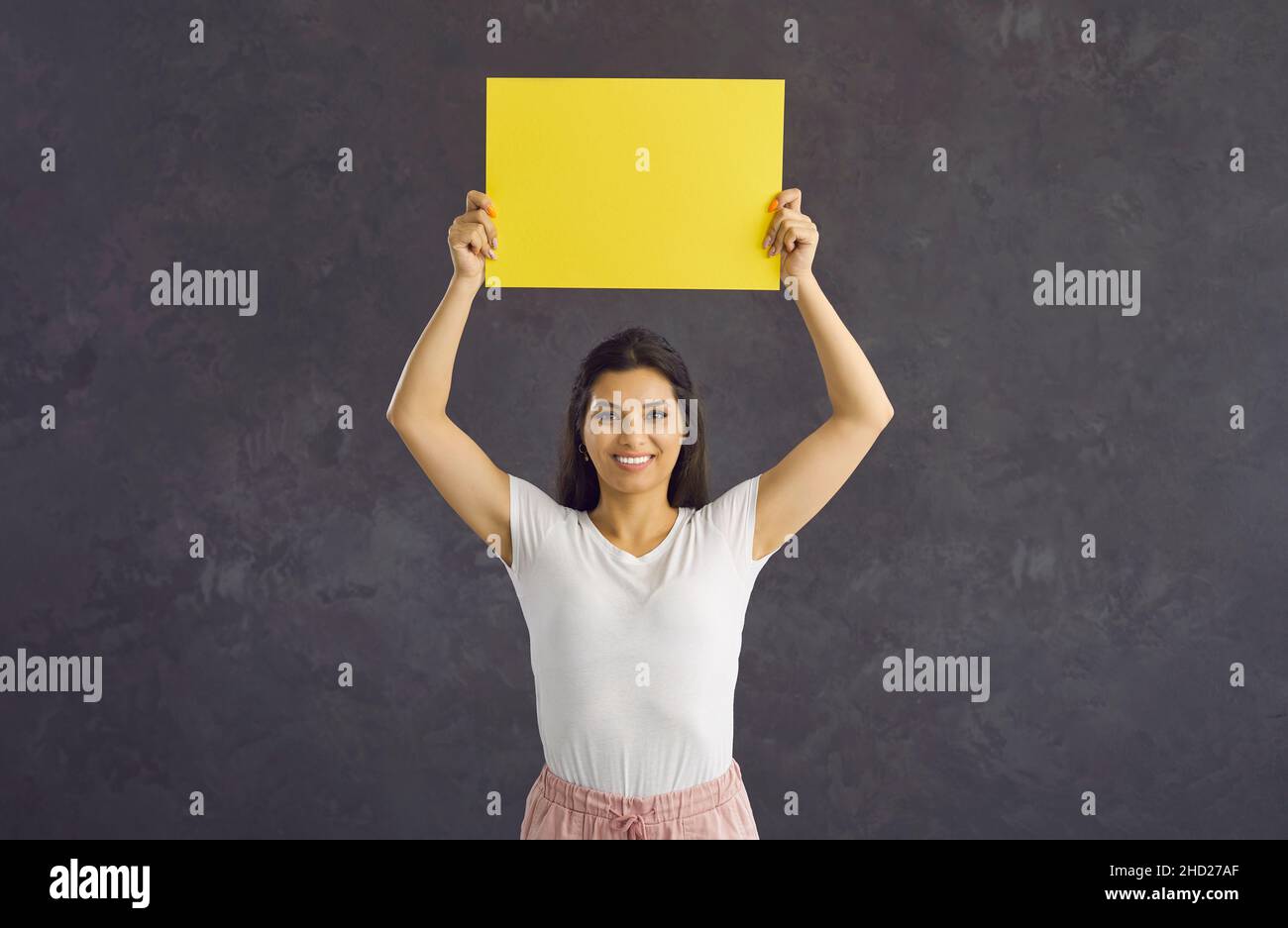 Positive joyful woman standing on gray background holding blank yellow sheet of paper for text. Stock Photo