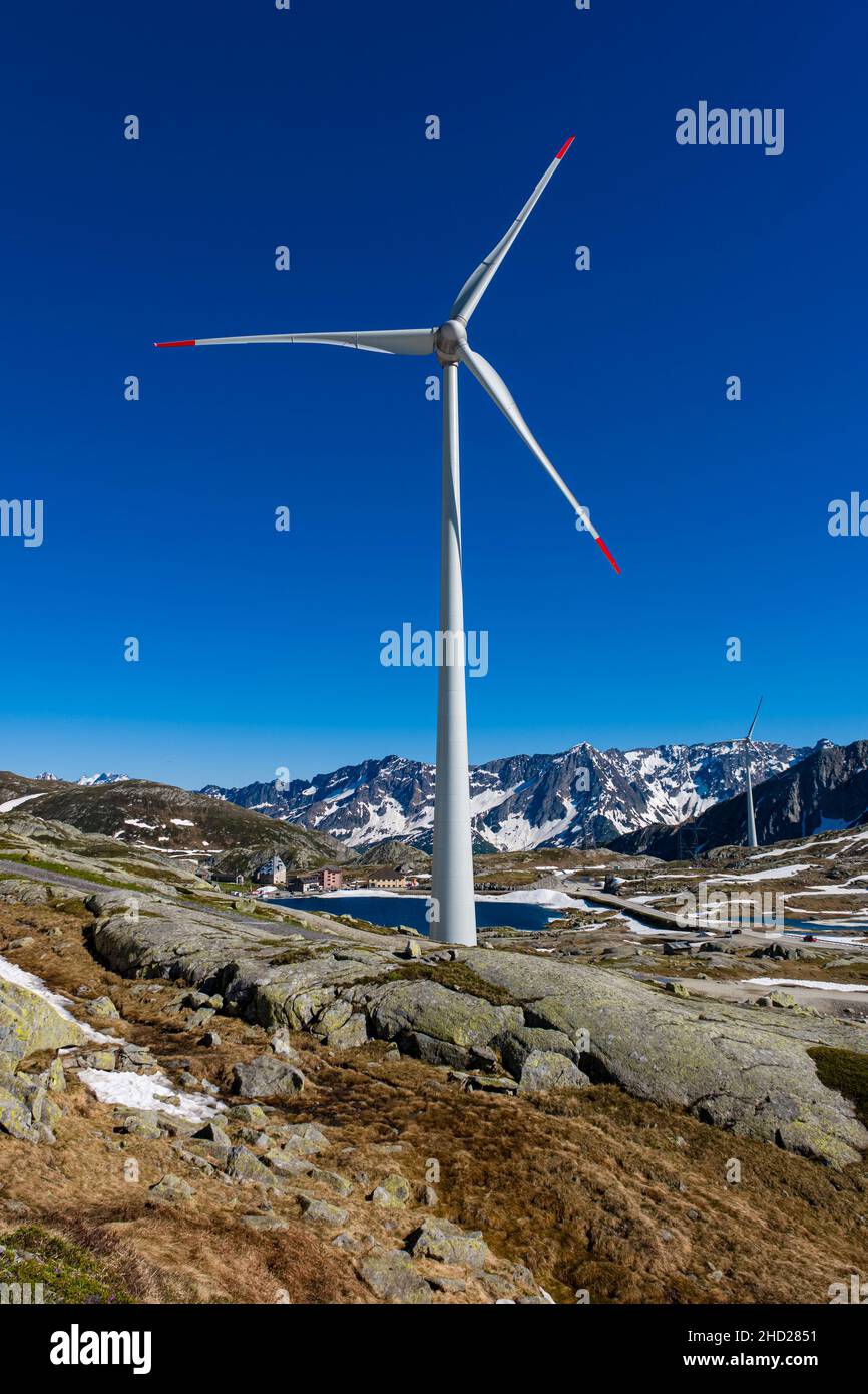 A wind turbine, seen across a lake, on top of Gotthard Pass at 2106 m, surrounding mountains covered in snow. Stock Photo