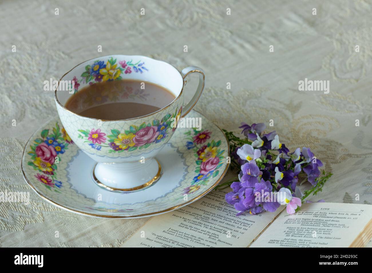 Cup of tea in a floral china cup with saucer on a patterned tablecloth with flowers and small book. Stock Photo