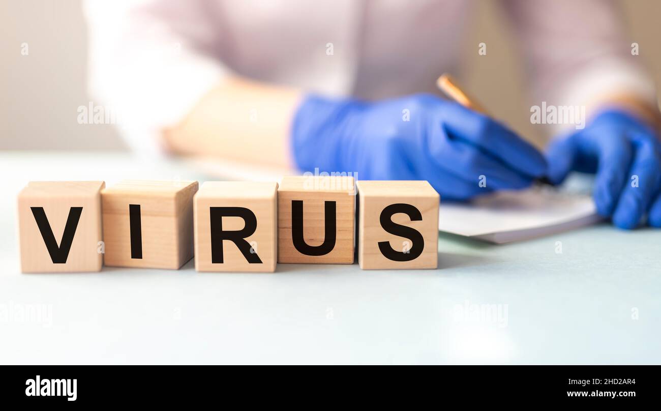 Word VIRUS on cube blocks and blurring hands of a female doctor in protective gloves writing smth Stock Photo