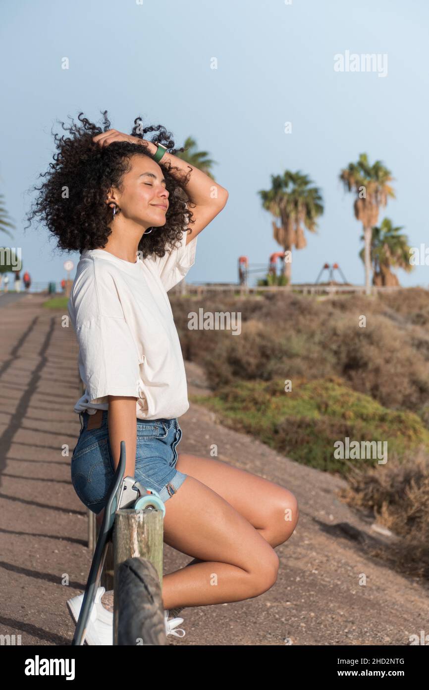 Young skater woman with afro hair sitting on a fence feeling the breeze and the sun on her face Stock Photo