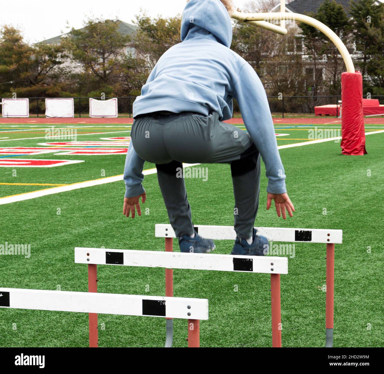 Rear view of a high school boy wearing a blue hoodie jumping over track hurdles on a green turf field during track practice. Stock Photo