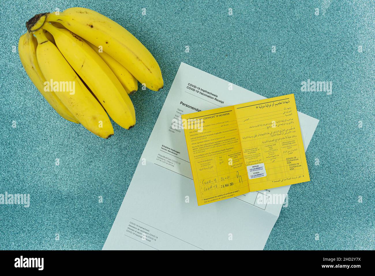 Yellow bananas and first documents and vaccine card with rubber stamp of covid-19 vaccine on turquoise background. Travel and covid-19 theme. Stock Photo