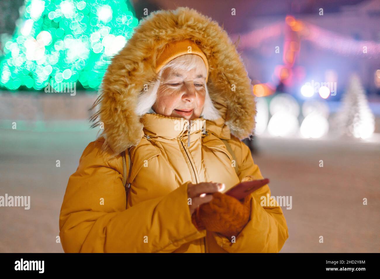 40s 50s woman in a yellow warm jacket using phone outdoors on street background decorated with Christmas lights at evening in winter. Communication Stock Photo