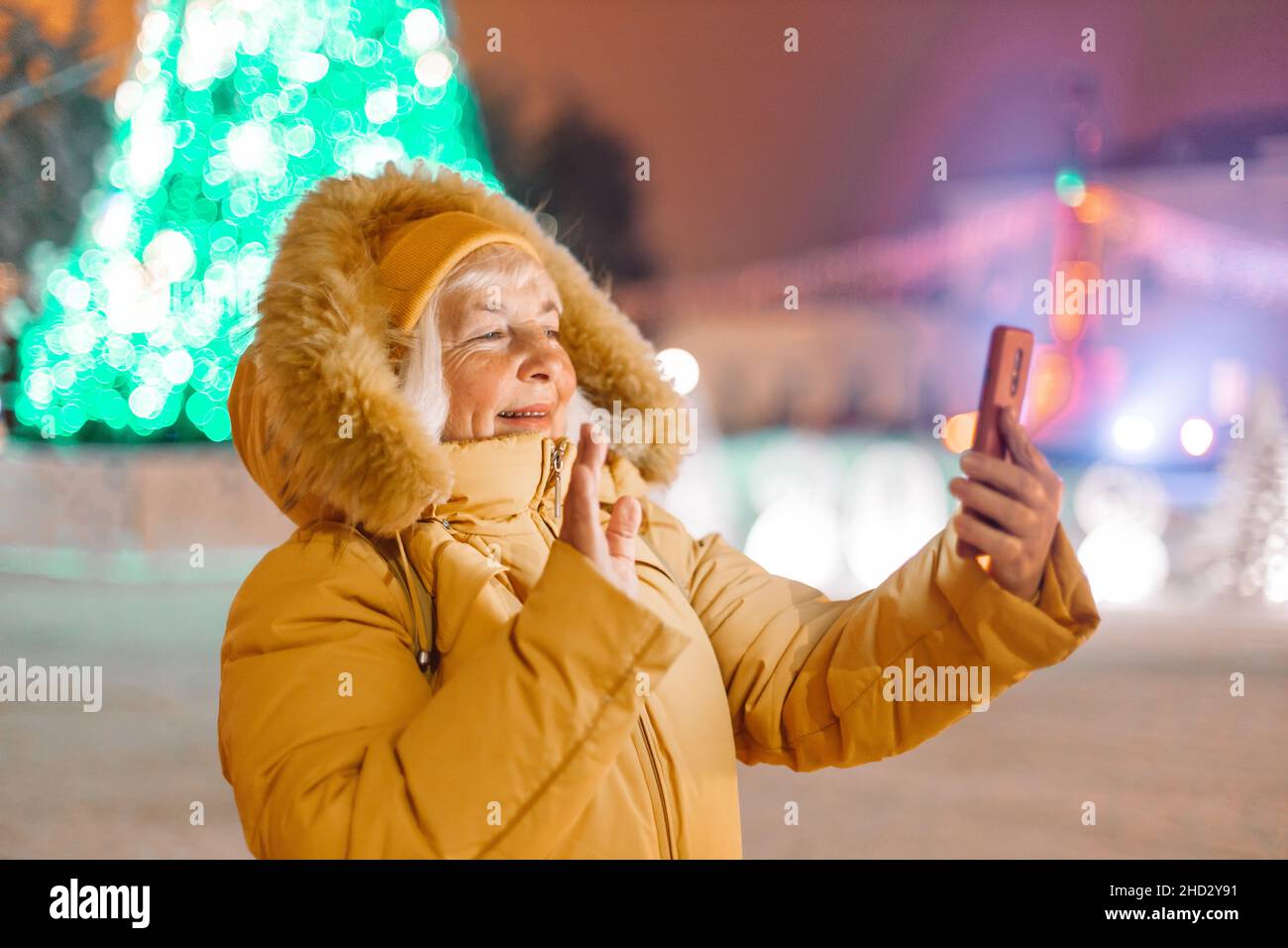 happy 50s blonde mature woman making selfie outdoors on street background decorated with Christmas lights at evening in winter during Christmas and Stock Photo