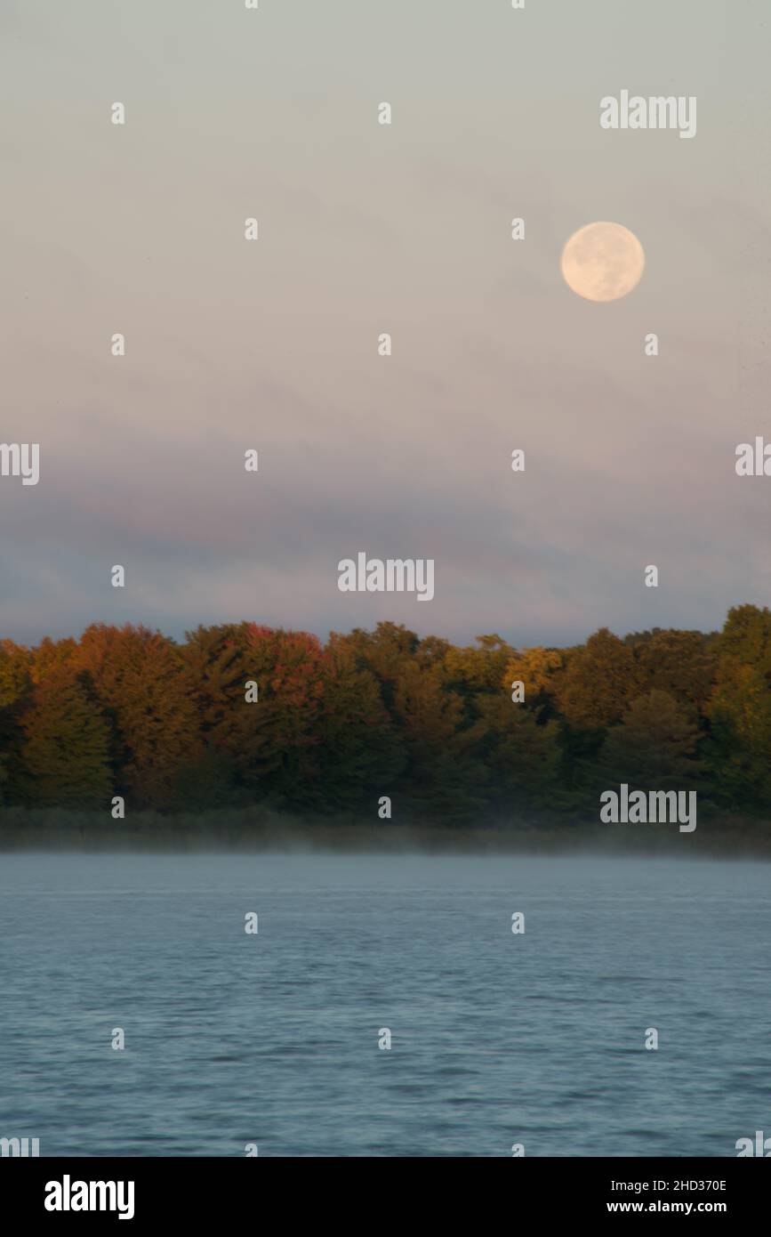 The full harvest moon through thin clouds over a lake with autumn colors on the far bank. Stock Photo