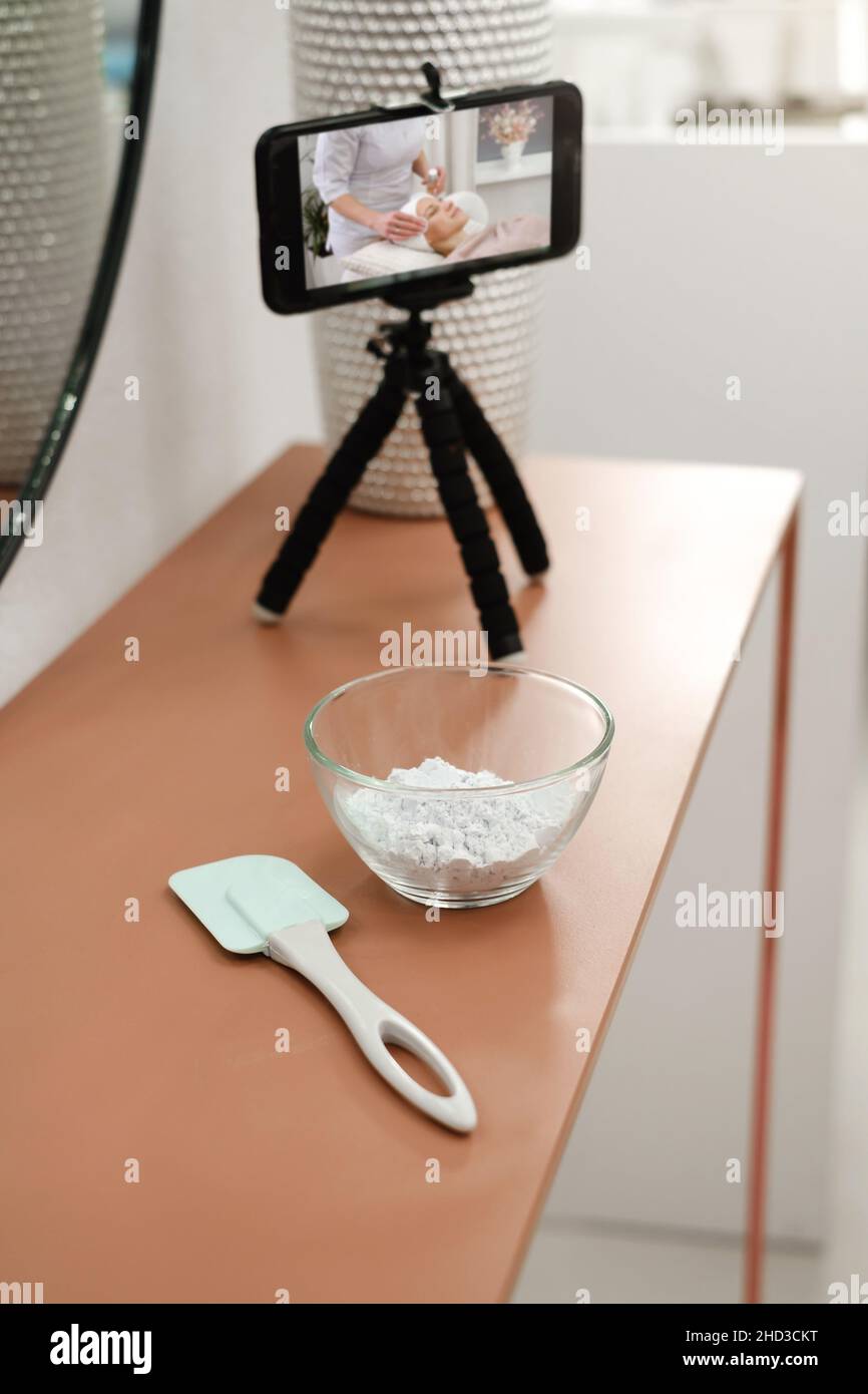 Beauty blogger streaming skin care therapy at beauty salon. Closeup of smartphone, powder for face mask on table. Cosmetician doing scrub. Lesson or Stock Photo
