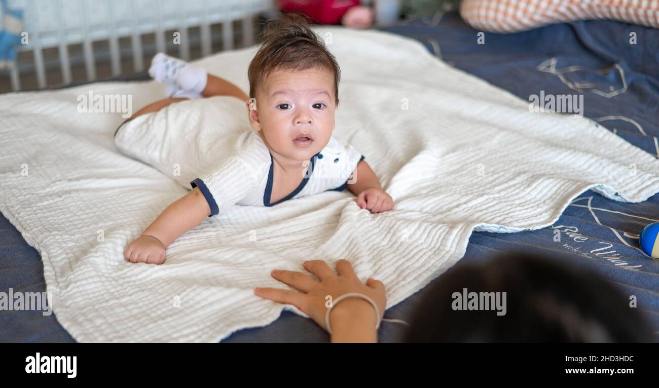 Mother playing with her 2 months old mixed race newborn baby boy lying on his tummy on the bed lifting head and playing Stock Photo