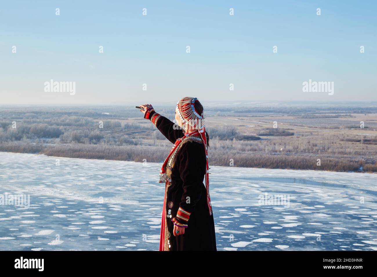 A young woman stands on the edge of a cliff with an outstretched hand with a jew's harp in winter dressed in a national Bashkir costume. Stock Photo