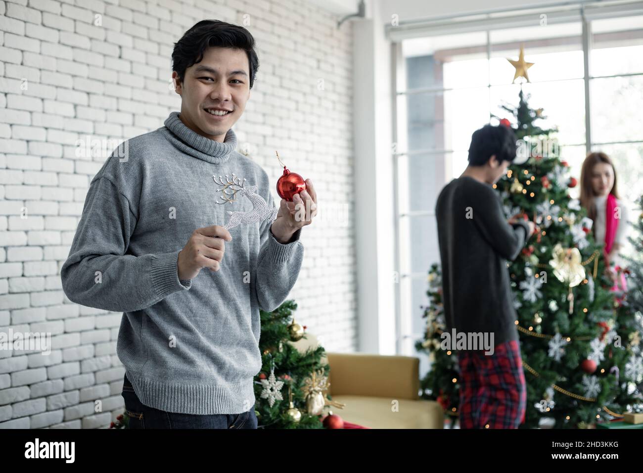 Young Asian man holds the toy to decorate the Christmas tree with smile on which has friends background. Celebration of new year at home. Merry Christ Stock Photo