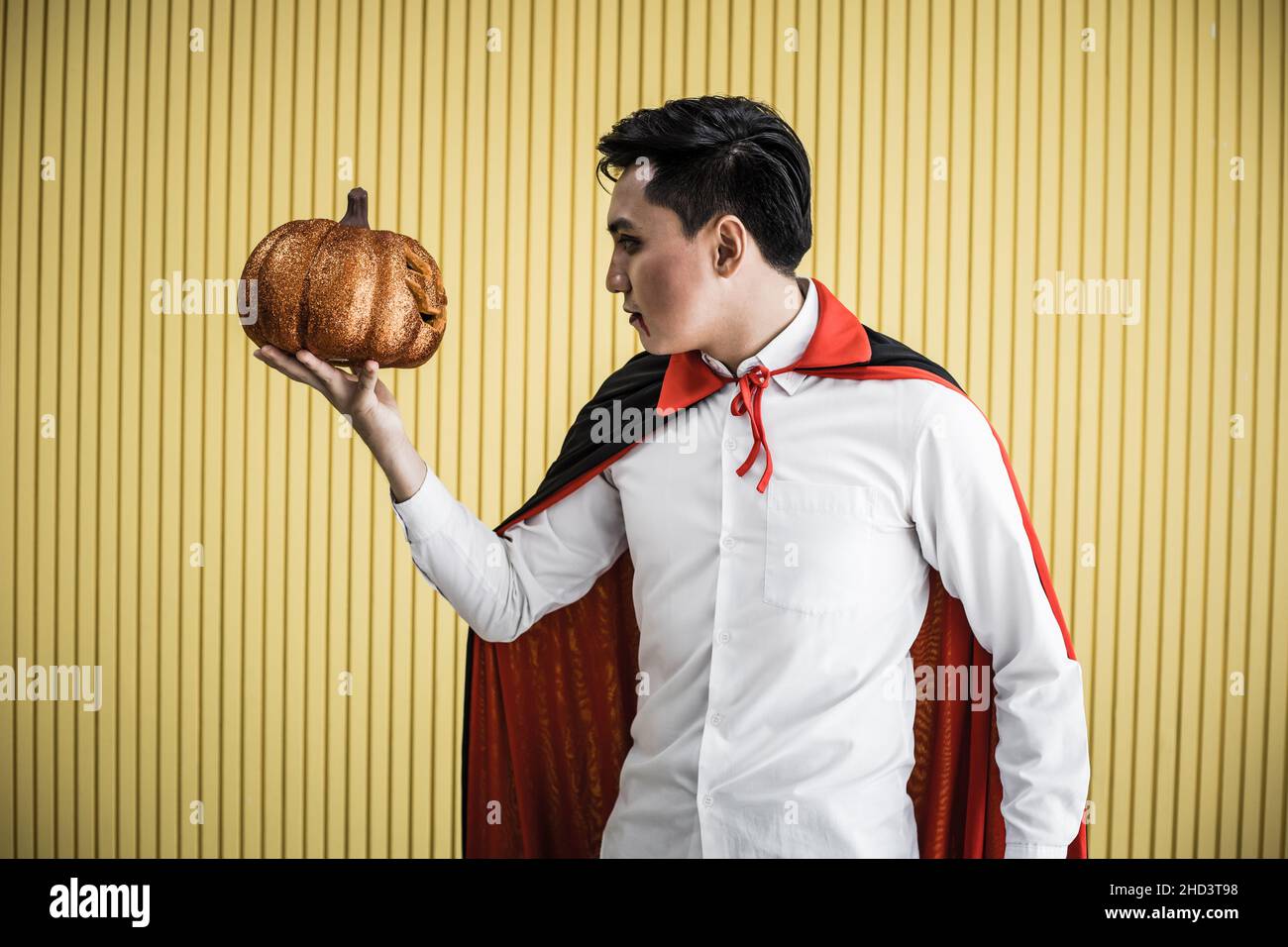 Halloween concept of young Asian man in costume dracula and hold Halloween orange pumpkin on yellow background. Portrait of teen man dressed up as dra Stock Photo