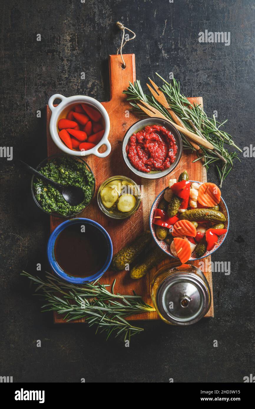 Bowls with various pickled vegetables snack on wooden cutting board with fresh herbs, pesto and homemade ketchup on dark rustic background. Top view Stock Photo