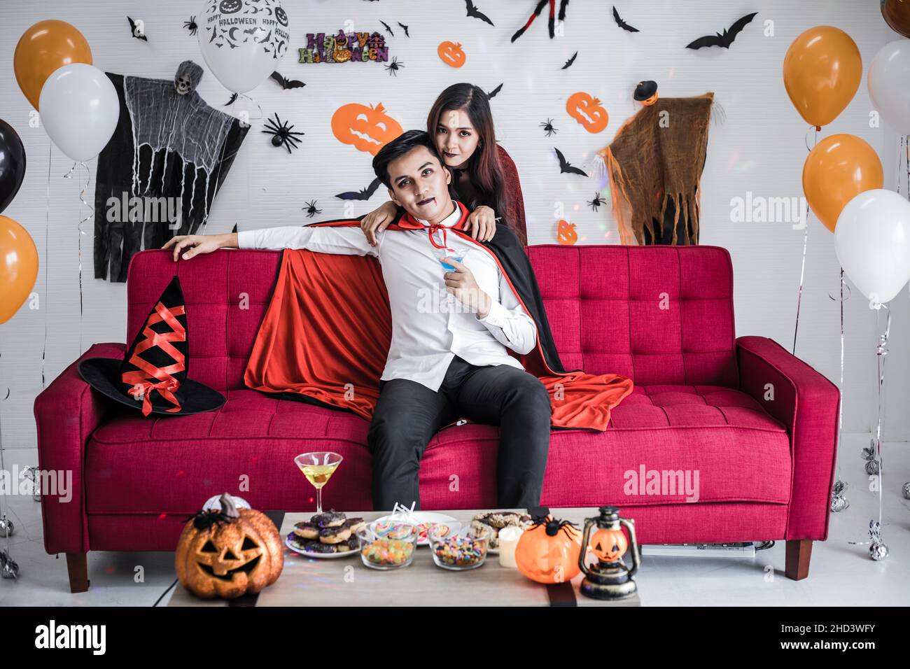 Young Asian couple in costume witch and dracula with celebrate Halloween party and drink wine together in Halloween festival on red sofa in the room a Stock Photo