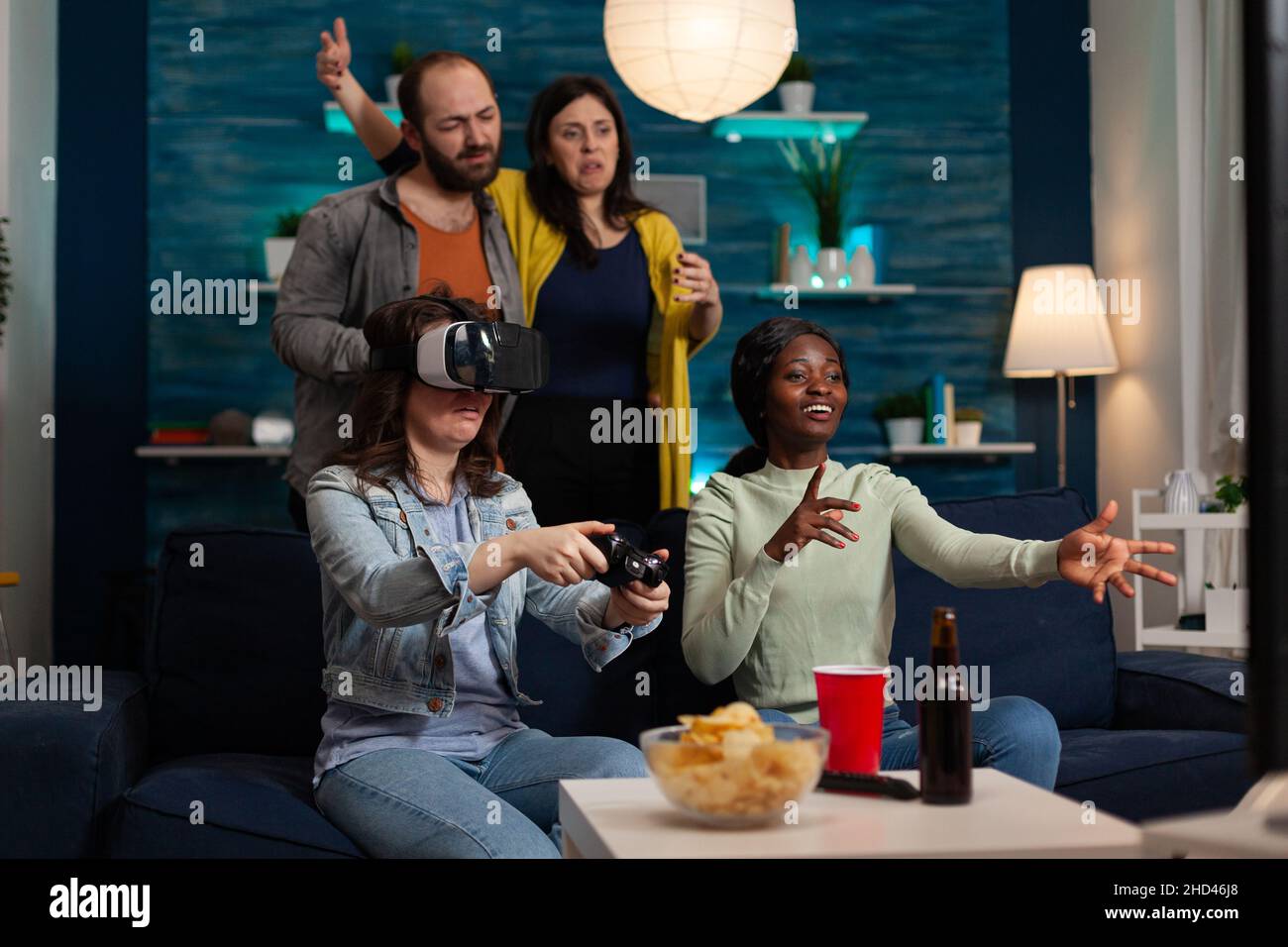 Gamer woman wearing virtual reality headset holding gaming controller playing videogames losing online competition. Group of multi-ethnic friends enjoying hanging out together making fun activity Stock Photo