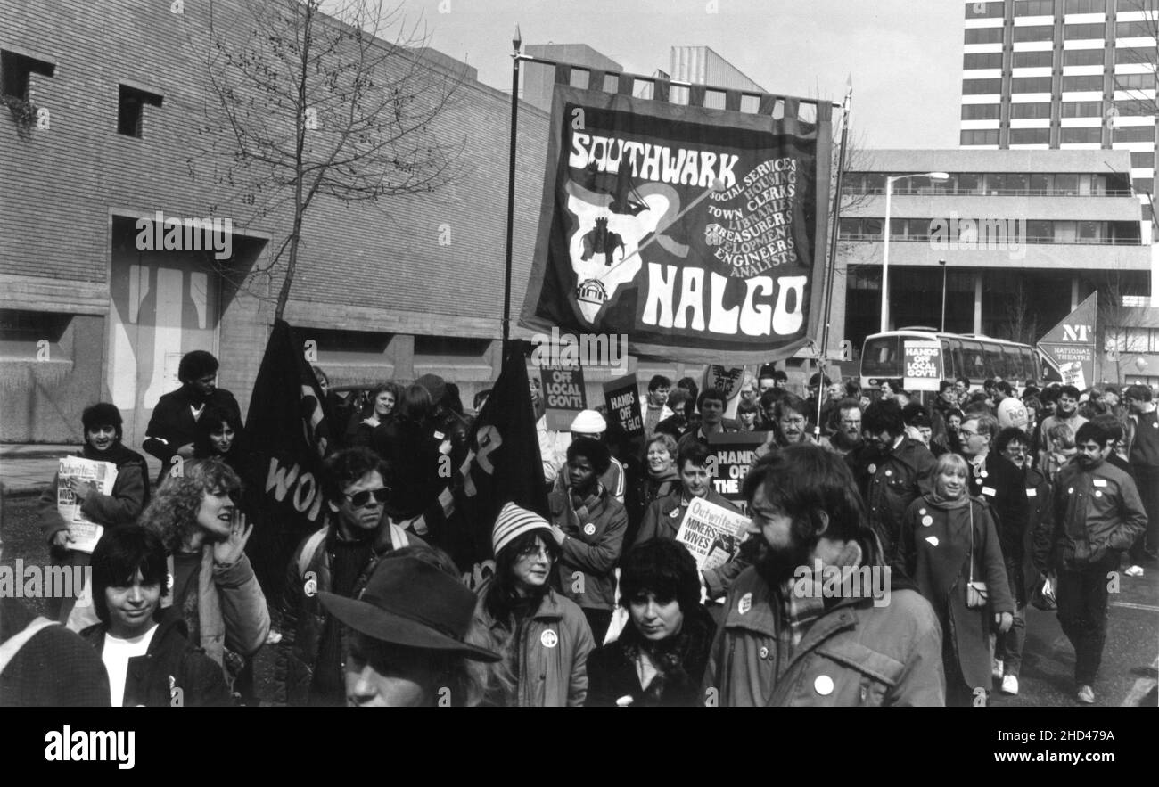 A 1984 demonstration march by members of the Southwark branch of the British trade union, NALGO (National and Local Government Officers' Association) protesting against the planned abolition of the GLC (Greater London Council). The photograph depicts the marchers passing by the National Theatre, Upper Ground, Southwark, London. The protestors are carrying the Southwark Branch NALGO banner, placards with the slogans, “Hands off Local Govt”, “Hands off The GLC” and also copies of the feminist newspaper ‘Outwrite’ bearing the headline, “Miners Wives – Media Lies”. Stock Photo