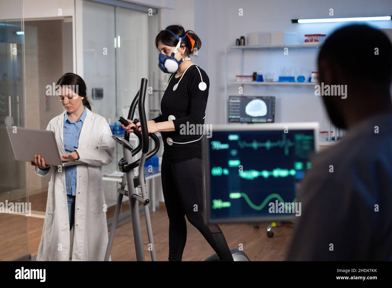 Athlete sportwoman with electrodes attached to her body running on cross trainer doing cardio exercise. Specialist researcher holding laptop analyzing endurance controling EGK data in modern lab Stock Photo