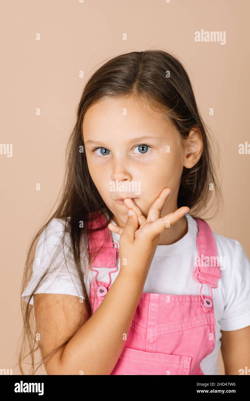 female kid biting fingernails with shining eyes looking at camera wearing bright pink jumpsuit and white t-shirt on beige background Stock Photo
