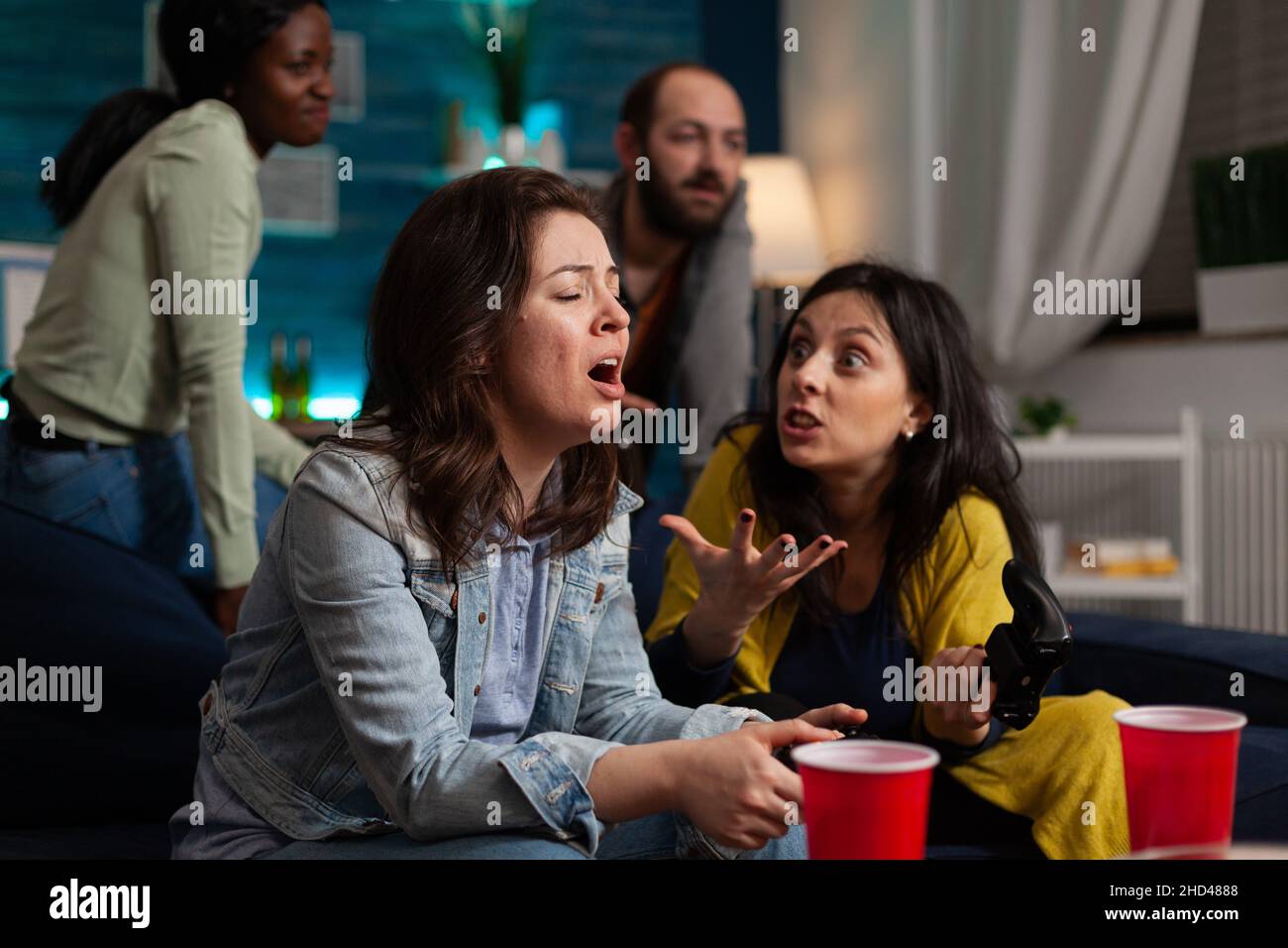 Nevours upset women sitting on sofa playing video games using gaming joystick losing online competition. Group of multi-ethnic friends hanging out together having fun during wekeend party at home Stock Photo