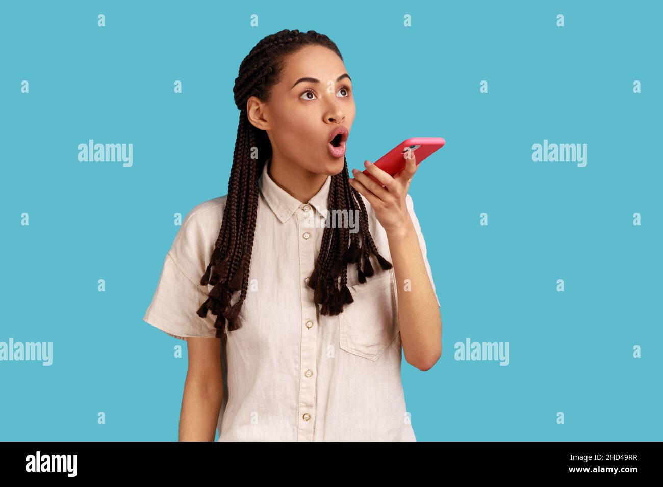 Surprised emotional woman with black dreadlocks uses voice for recognition app on modern cell phone, recording message, wearing white shirt. Indoor studio shot isolated on blue background. Stock Photo