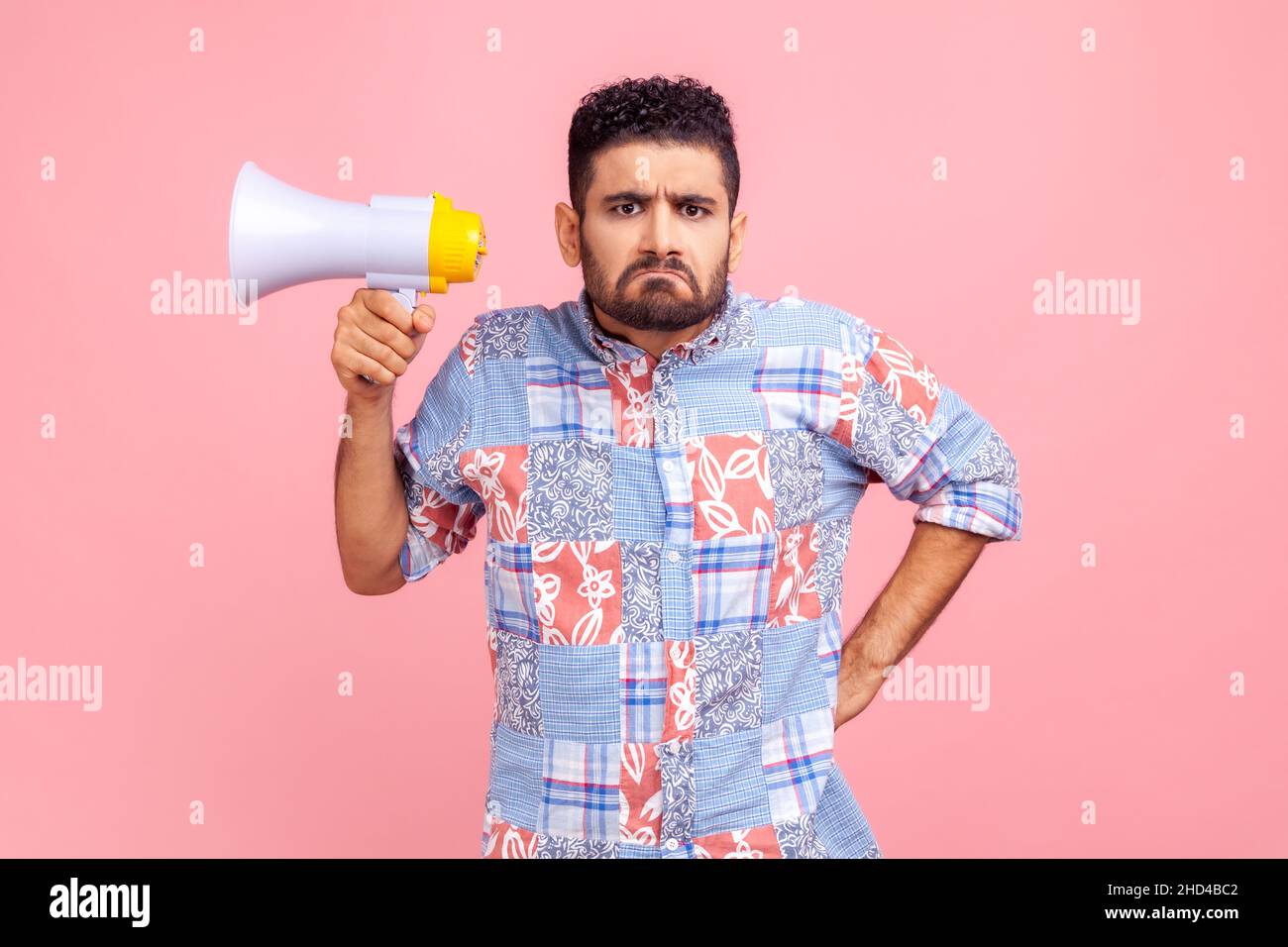 Portrait of strict serious upset man with frowning face holding megaphone looking at camera with sad expression, keeping hand on hip. Indoor studio shot isolated on pink background. Stock Photo
