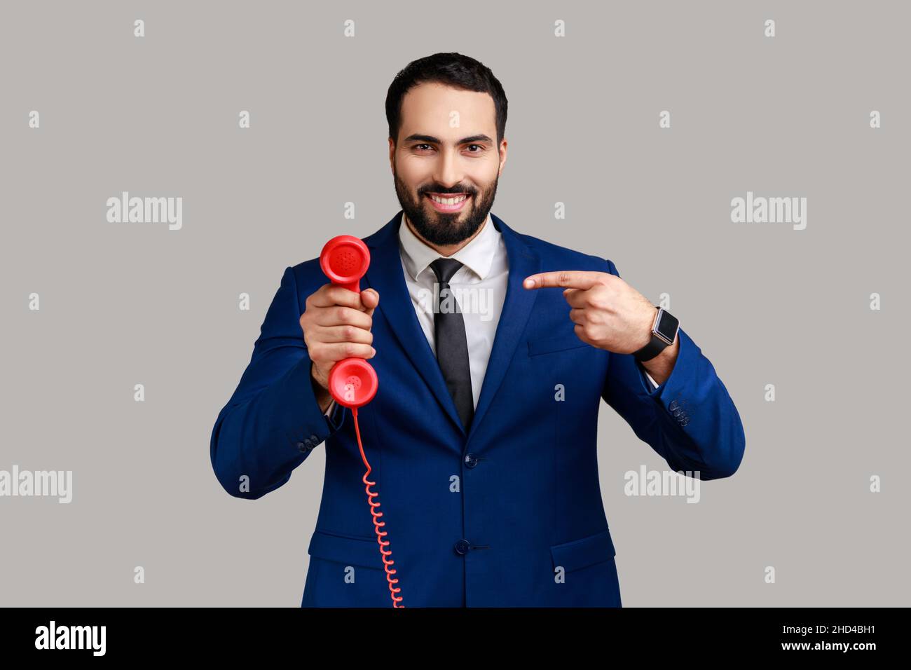 Bearded man pointing finger at handset of red vintage landline phone, interested in retro devices, answering calls, wearing official style suit. Indoor studio shot isolated on gray background. Stock Photo