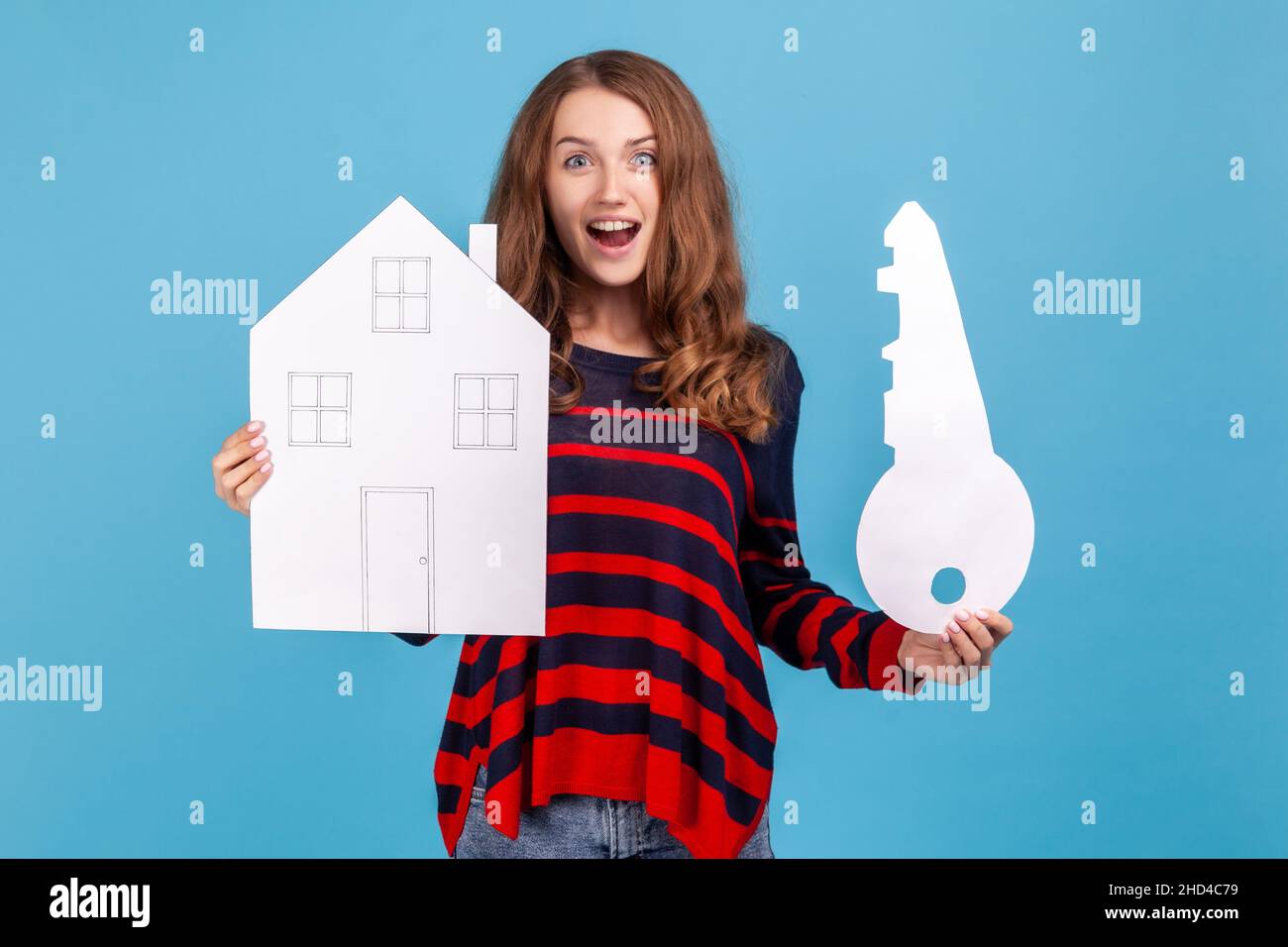 Surprised astonished woman wearing striped casual style sweater, holding paper house and big key, real estate purchase, rental services, mortgage. Indoor studio shot isolated on blue background. Stock Photo