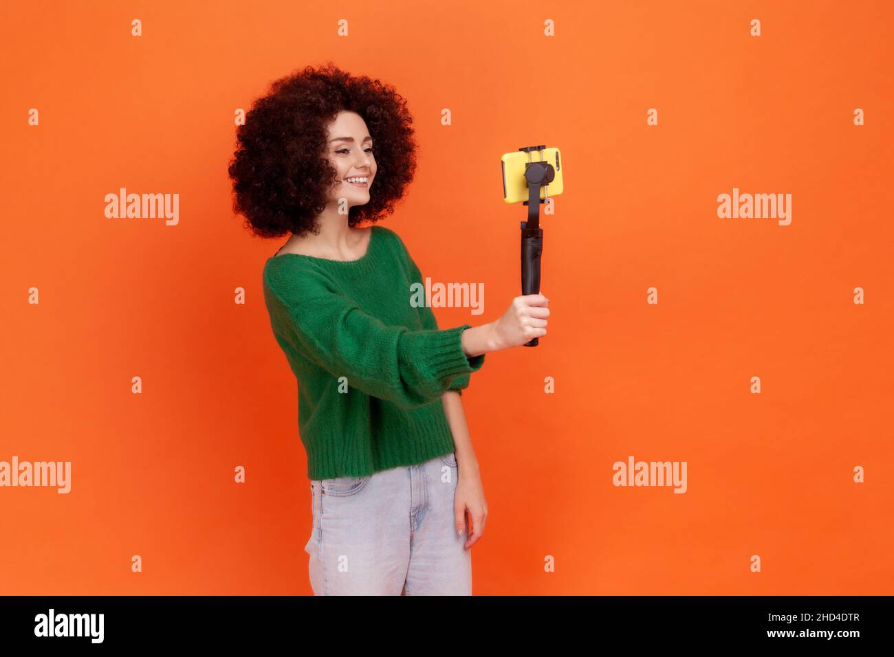 Side view portrait of woman with Afro hairstyle wearing green casual style sweater talking to her followers, online conversation, livestream. Indoor studio shot isolated on orange background. Stock Photo