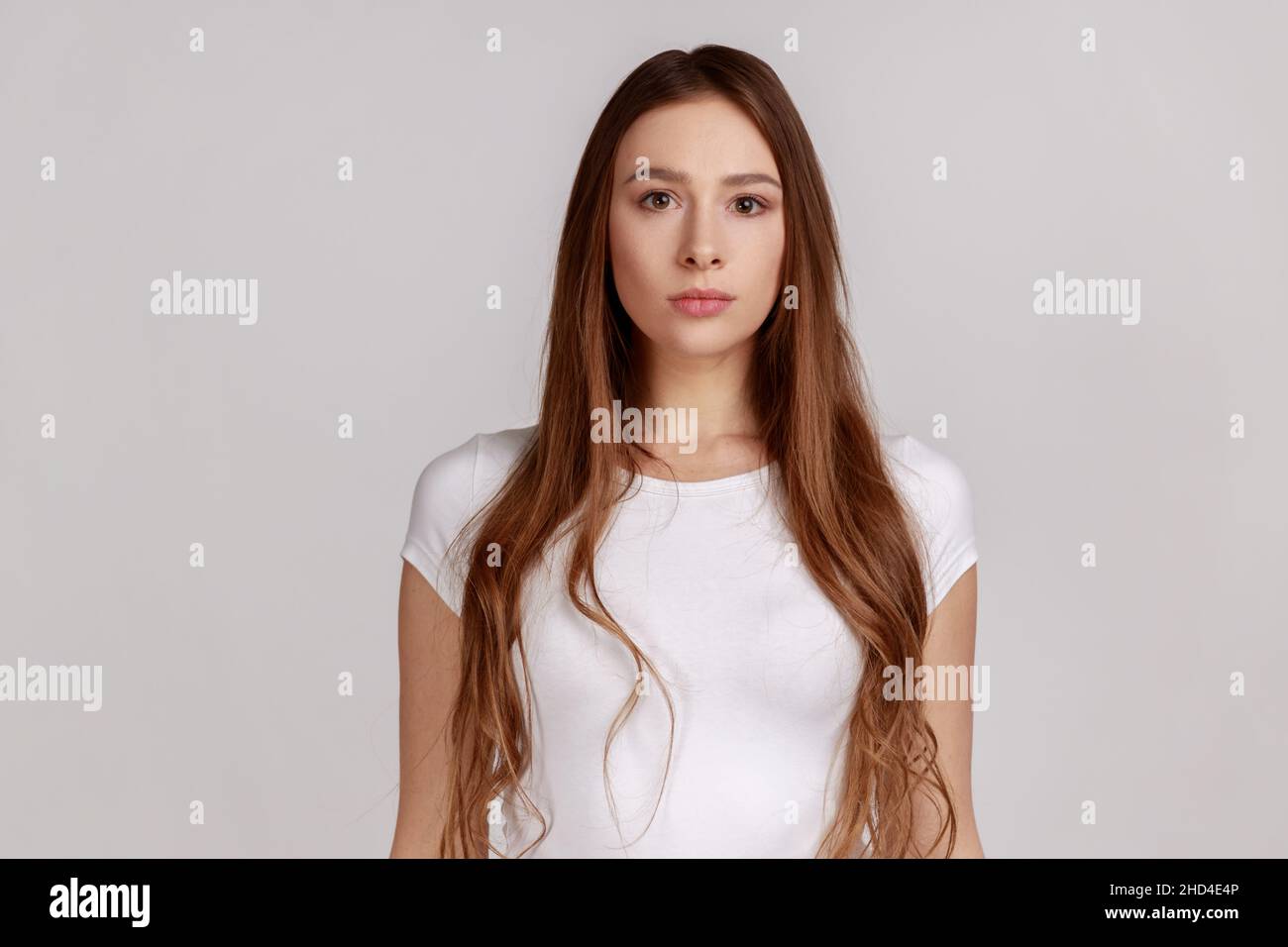 Portrait of strict attractive woman standing looking at camera, feels confident focused self-assured, expressing seriousness, wearing white T-shirt. Indoor studio shot isolated on gray background. Stock Photo