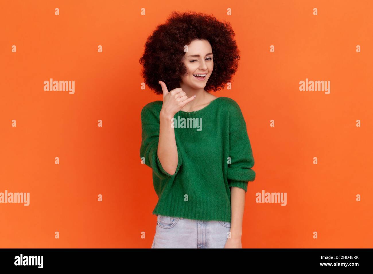 Woman with Afro hairstyle holding fingers shaped like telephone near head, communicating by phone, looking at camera with playful smile. Indoor studio shot isolated on orange background. Stock Photo