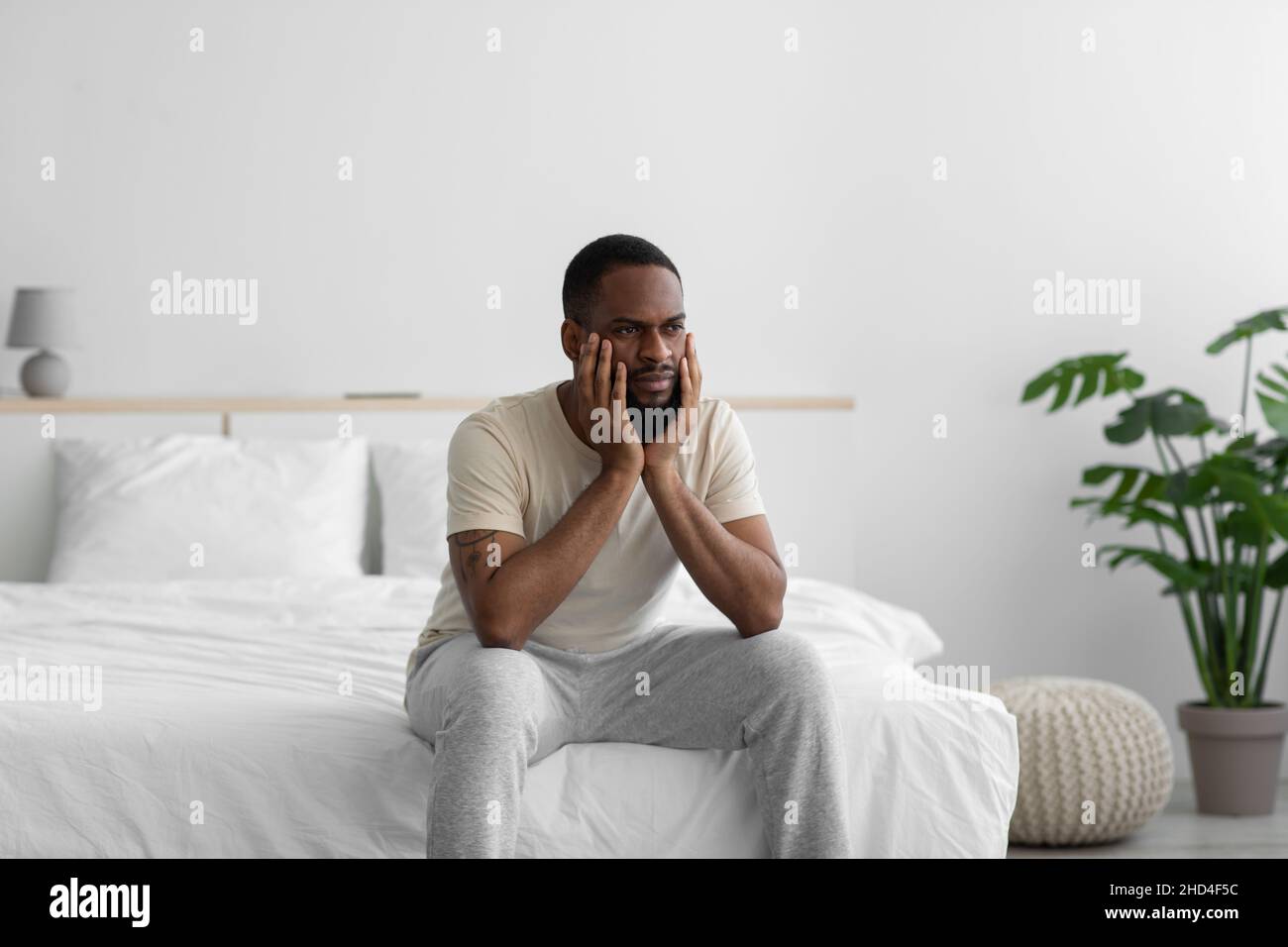 Sad pensive millennial african american guy in domestic clothes ponders solution to problem in white minimalist bedroom Stock Photo