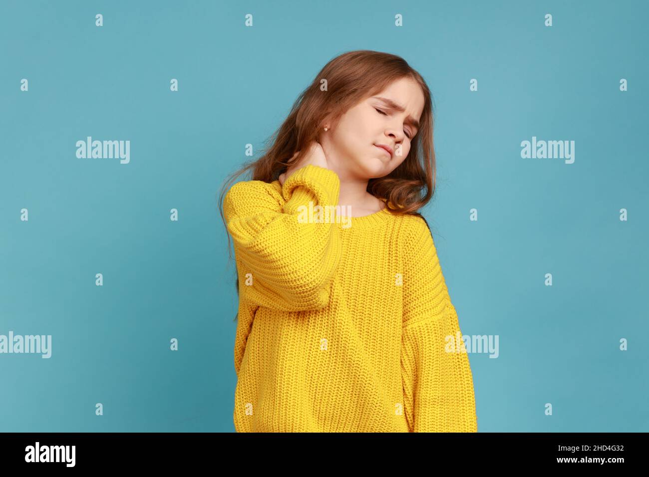 Portrait of little girl massaging painful back, feel discomfort in neck, child sedentary lifestyle, wearing yellow casual style sweater. Indoor studio shot isolated on blue background. Stock Photo