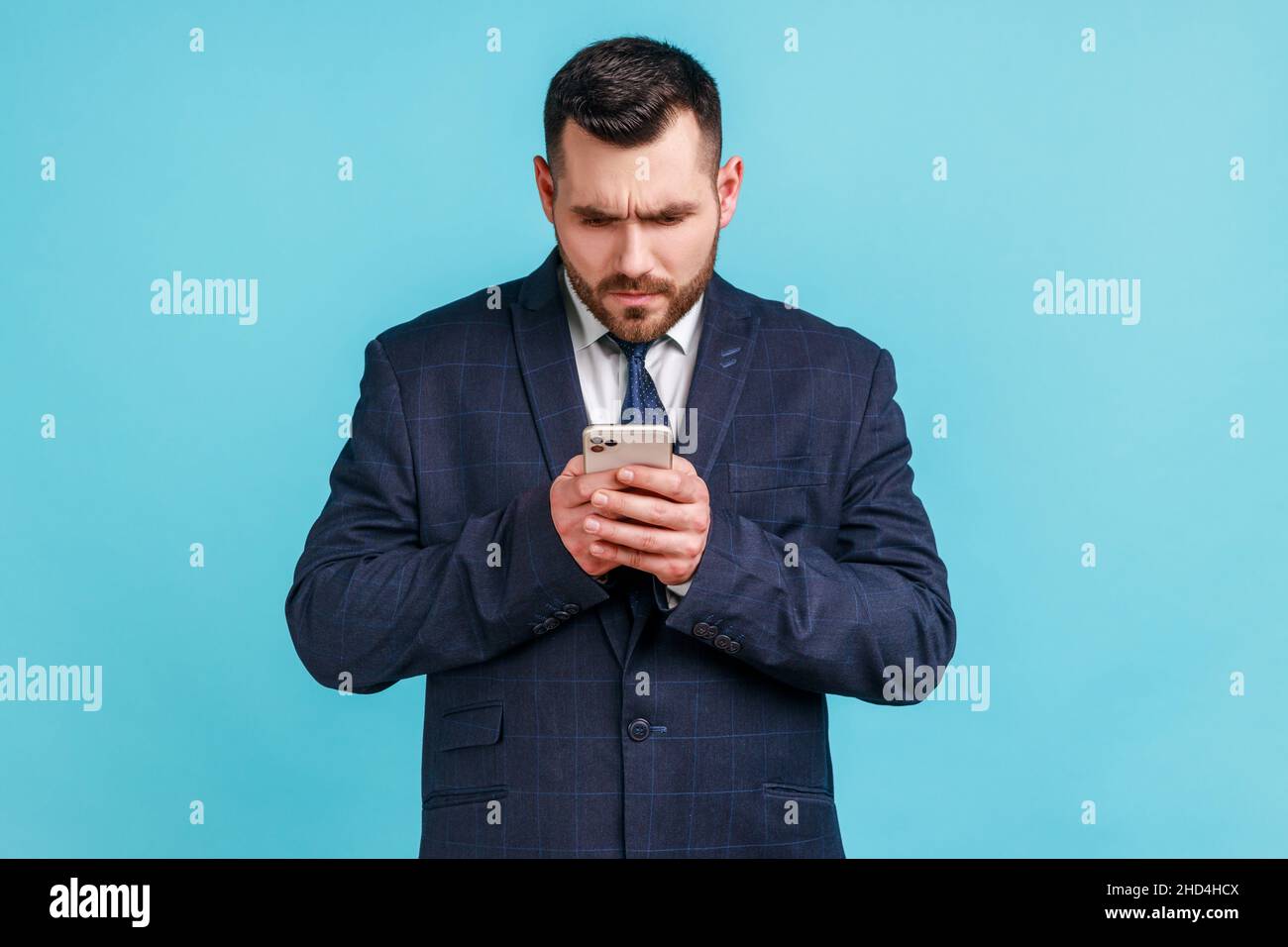 Serious bearded man wearing official style suit texting message in social media on cell phone, using mobile network services, chatting online. Indoor studio shot isolated on blue background. Stock Photo