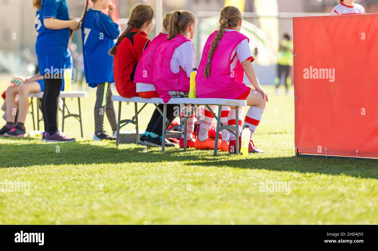 School girls sitting on football bench and watching tournament game. Female youth soccer team playing competition game on summer day. Female football Stock Photo