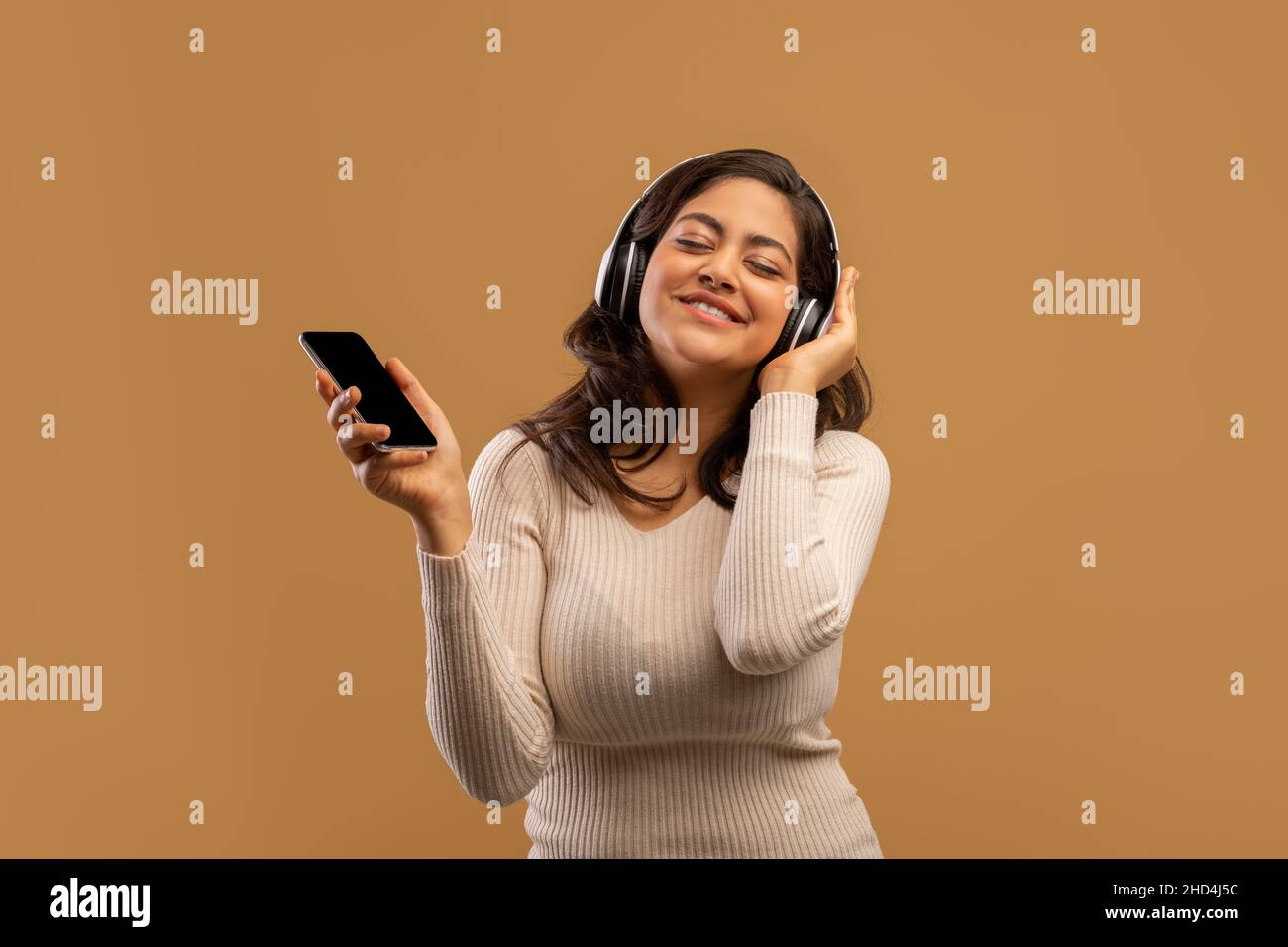 Playful arab lady with headphones and smartphone listening to music and enjoying cool playlist, beige background Stock Photo