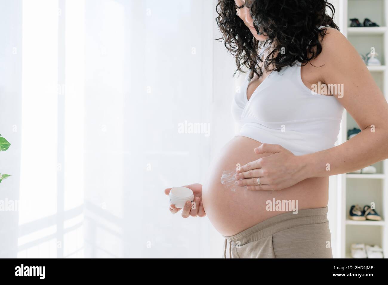 Young pretty pregnant woman with curly hair putting moisturizer on her tummy to prevent stretch marks in pregnancy.White background. copy space Stock Photo