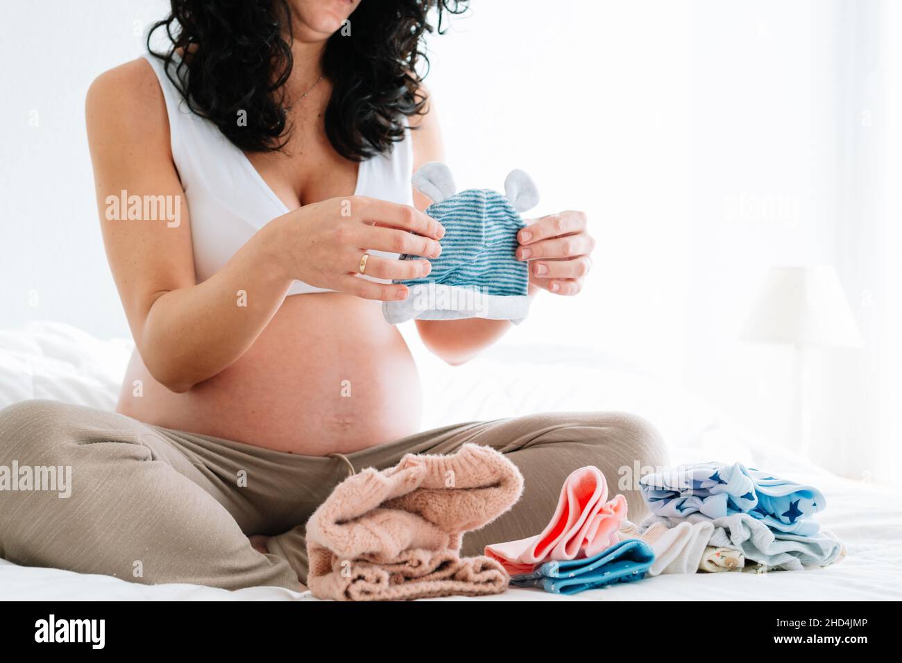 young pregnant woman with curly hair sitting on the bed looking at a little hat for her future newborn baby. folded baby clothes on the bed Stock Photo