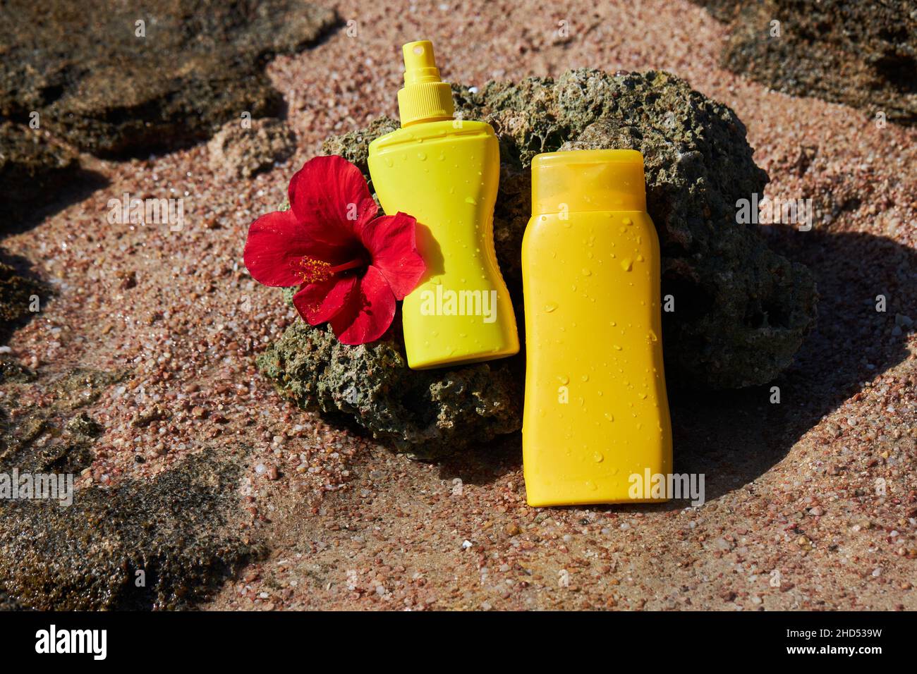 Yellow sunscreen cream bottles for skin protection with a red Hibiscus flower on a stone at the sea background. Beautiful water drops of sea waves Stock Photo