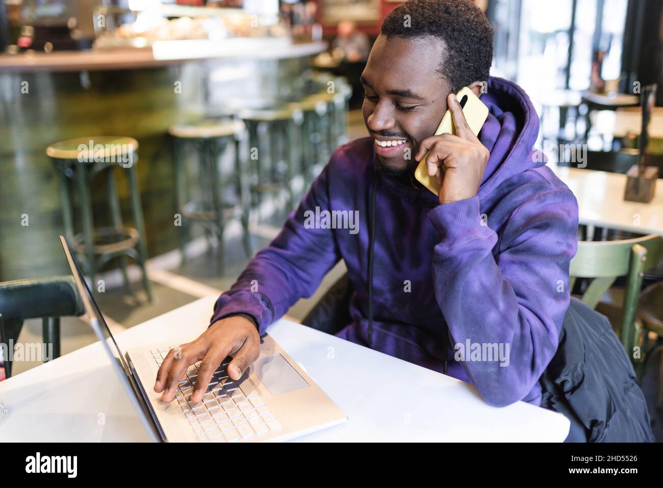 High angle of positive African American male freelancer having conversation on mobile phone while doing online project on netbook in cafe Stock Photo