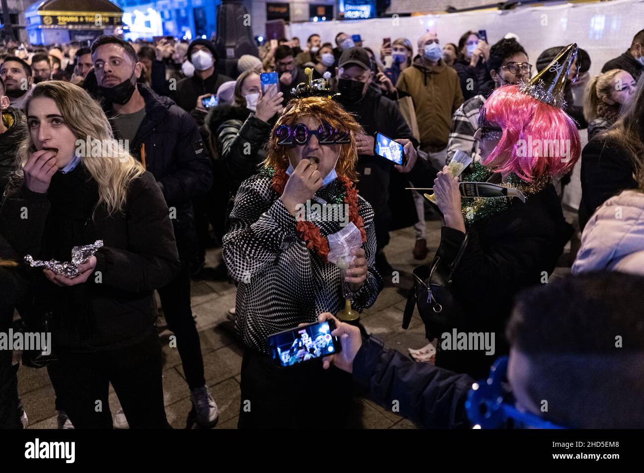 Madrid, Spain. 31st Dec, 2021. A man wearing a wig and 2022 googles is seen eating grapes in la Puerta de Sol during the preuvas celebrations in Madrid.Every year people gather in this Puerta del Sol in Madrid to take advantage of the New Year's Eve bells to celebrate the start of the new year in what is called the Preuvas celebration. (Credit Image: © Cristobal Ospina/SOPA Images via ZUMA Press Wire) Stock Photo