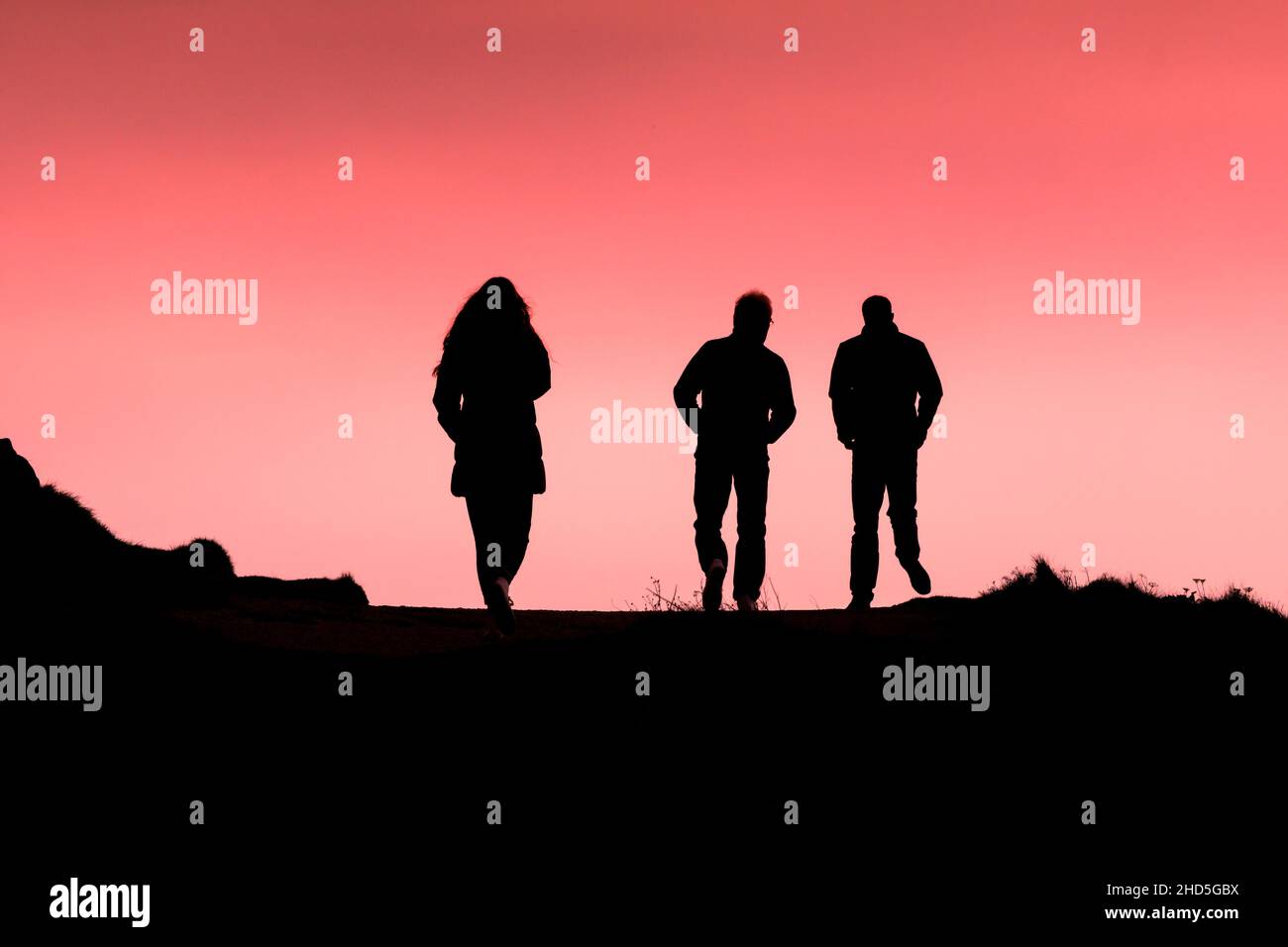 Three people walking along the coast path silhouetted against a colourful sky at the end of the day in Newquay in Cornwall. Stock Photo