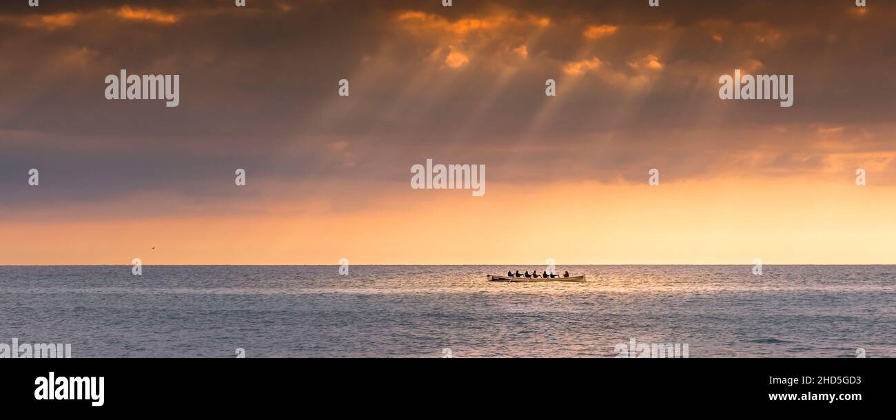 A panoramic image of the 200 year old Cornish Pilot Gig Dove being rowed as the sun sets over Fistral Bay in Newquay in Cornwall. Stock Photo