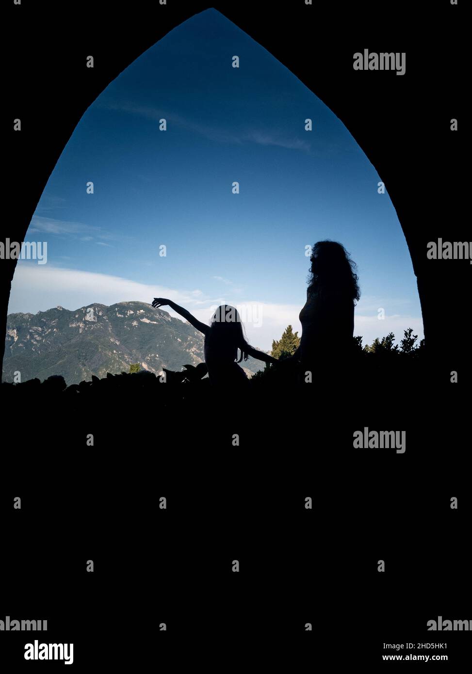 Silhouettes of female with a girl under an arched structure and rocky mountains in background Stock Photo