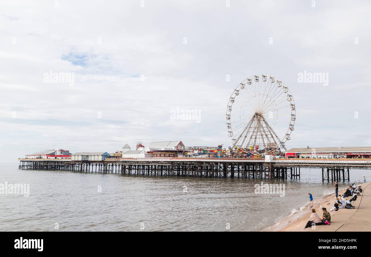 Tourists sit on the steps on Blackpool seafront watching the ferris wheel rotating on Central Pier. Stock Photo